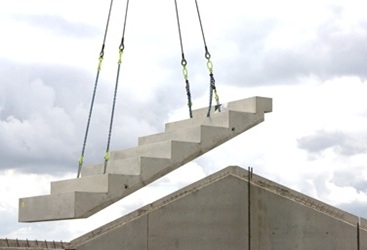Eine massive Betontreppe wird mithilfe von Seilen und einem Kran auf ein Gebäude gehoben. Die Treppe ist als Fertigteil vorgefertigt und wird auf der Baustelle präzise eingesetzt. Der Himmel ist bewölkt.

                                  
